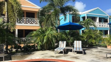 Zona de piscina con sillones de descanso y sombrillas, rodeada de palmeras y edificios de dos pisos con balcones.