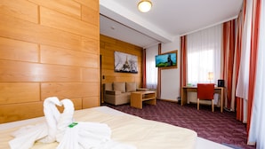 Habitación de hotel con cabecera de madera, cama con toallas blancas, sofá, escritorio con silla, televisor y ventana con cortinas.