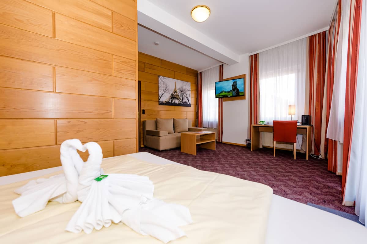 A hotel room with a wooden headboard, a bed with white towels, a sofa, a desk with a chair, a television, and a window with curtains.