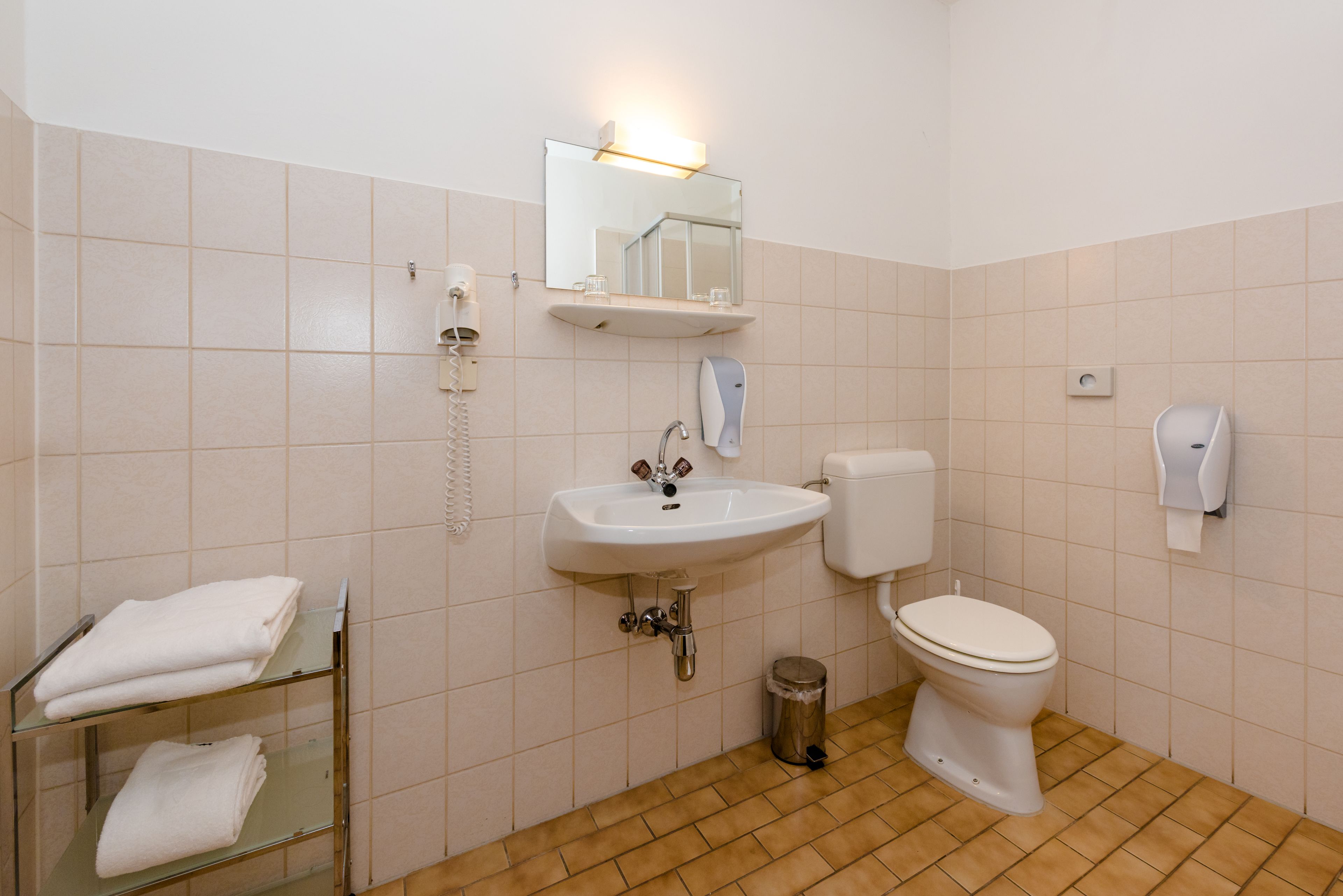Un baño con lavamanos, inodoro y espejo. Hay toallas en un estante y un cubo de basura.