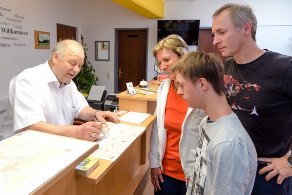 A reception desk with a man signing a document, a woman observing, and a child standing nearby. - Stadthotel Schwerterbräu (Judenburg)