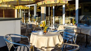Un área de comedor al aire libre con mesas y sillas, cubierta por sombrillas amarillas con la marca 'RAUCH', y una estructura de madera al fondo.