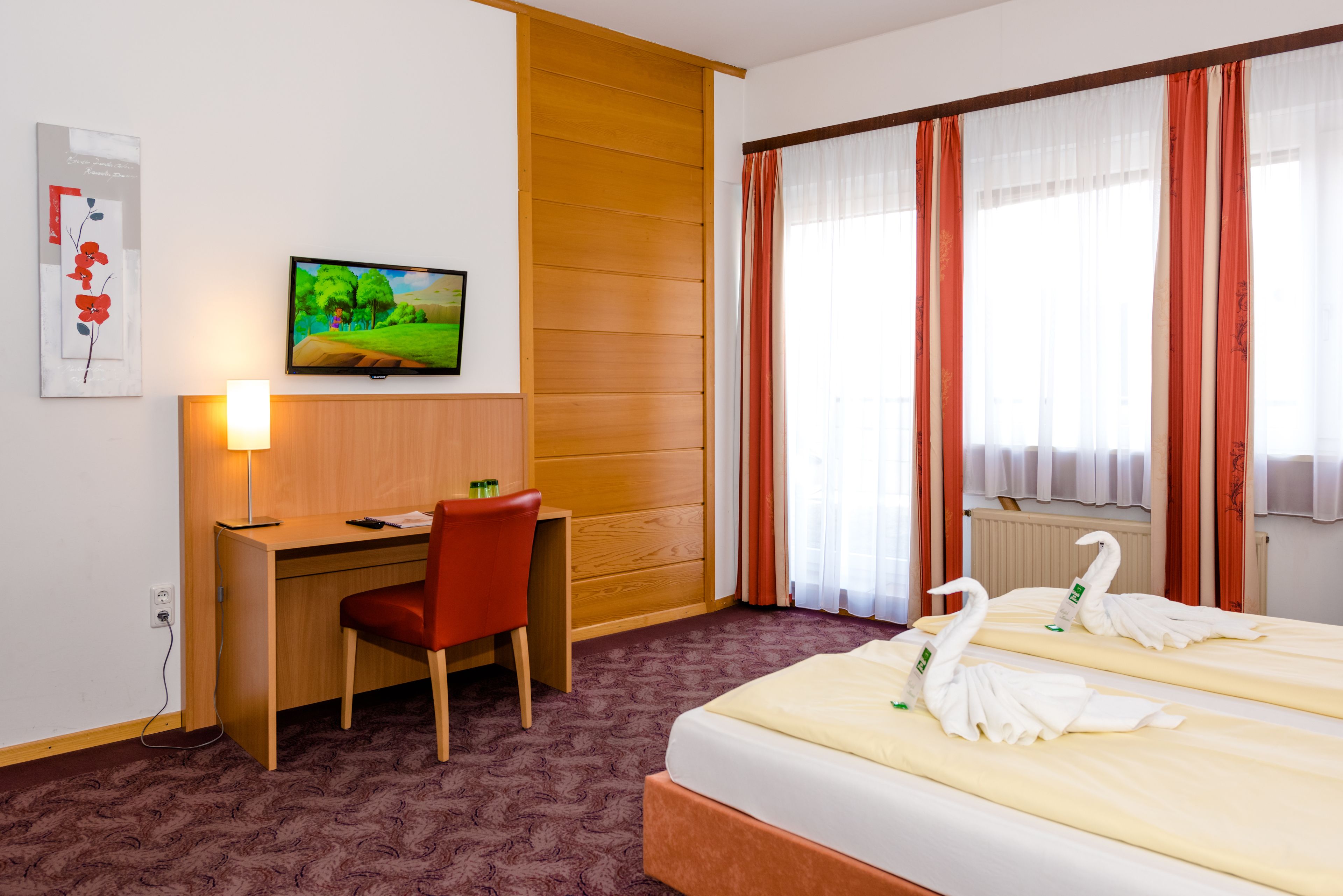 A hotel room with a wooden desk, a red chair, a bed with white towels shaped like swans, a television on the wall, and a window with red curtains.