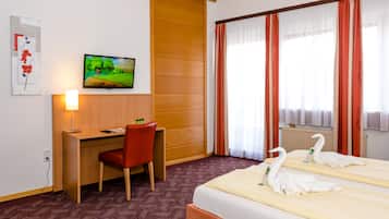 A hotel room with a wooden desk, a red chair, a bed with white towels shaped like swans, a television on the wall, and a window with red curtains.