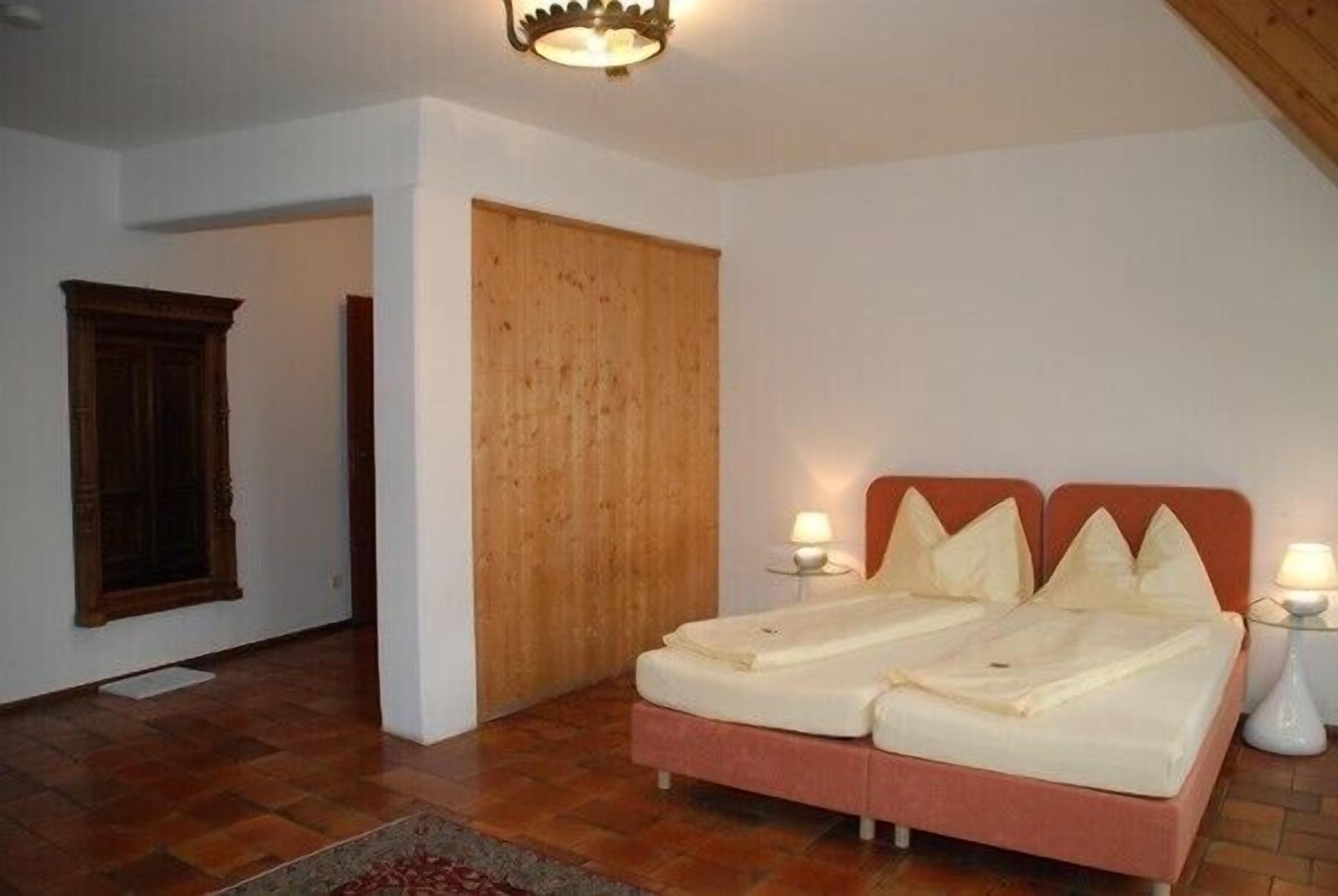 A bedroom with a double bed, two bedside lamps, a wooden door, and a window with a wooden frame.