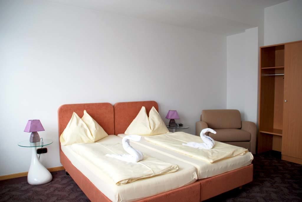 A hotel room with a double bed, two pillows, a glass table with a lamp, a chair, and a wardrobe.