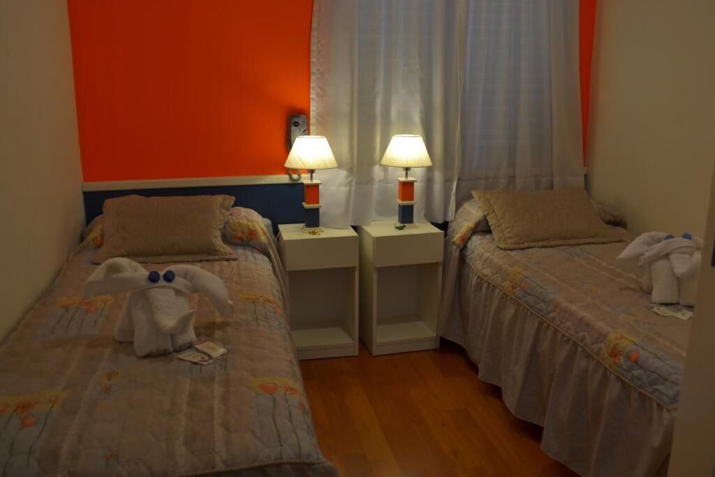 Una habitación de hotel con dos camas individuales, cada una con una mesita de noche y una lámpara. El piso es de madera y las paredes son anaranjadas.