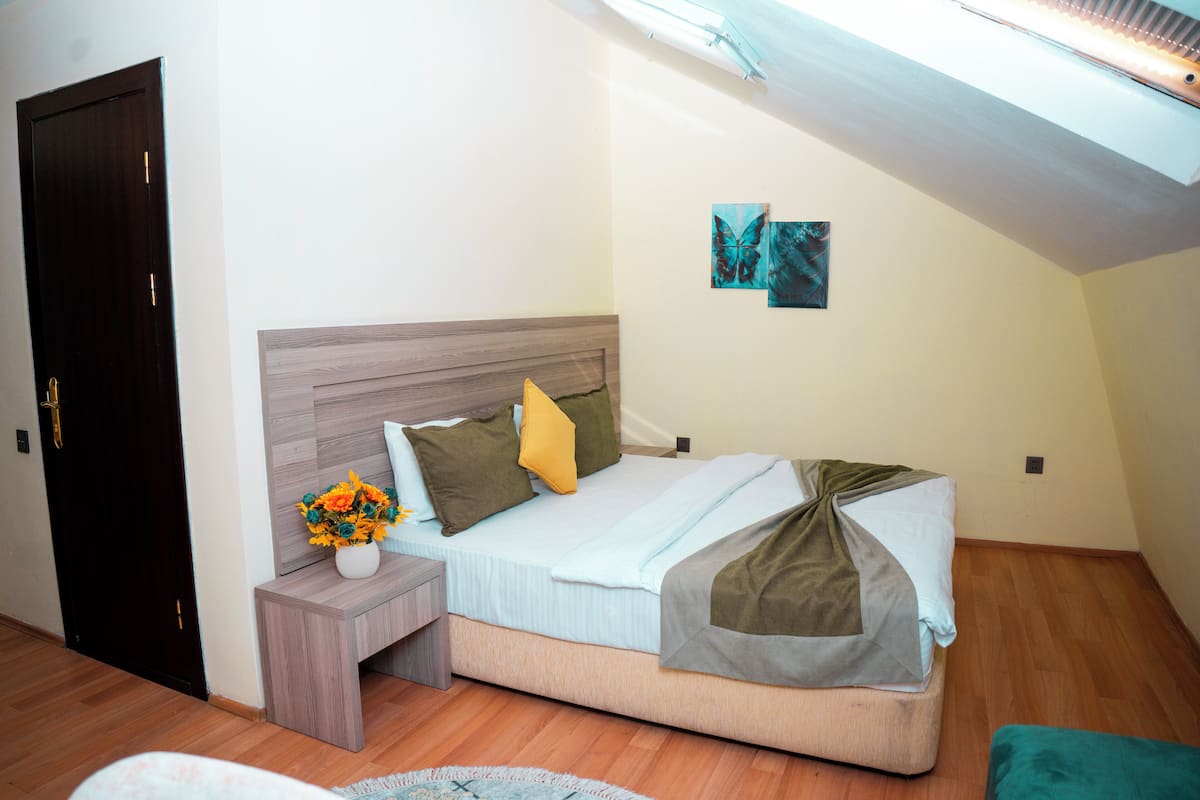 A bedroom with a wooden headboard, a bed with a light blue bedspread, a green throw blanket, and a yellow pillow. There is a nightstand with a vase of flowers and a small table beside the bed.