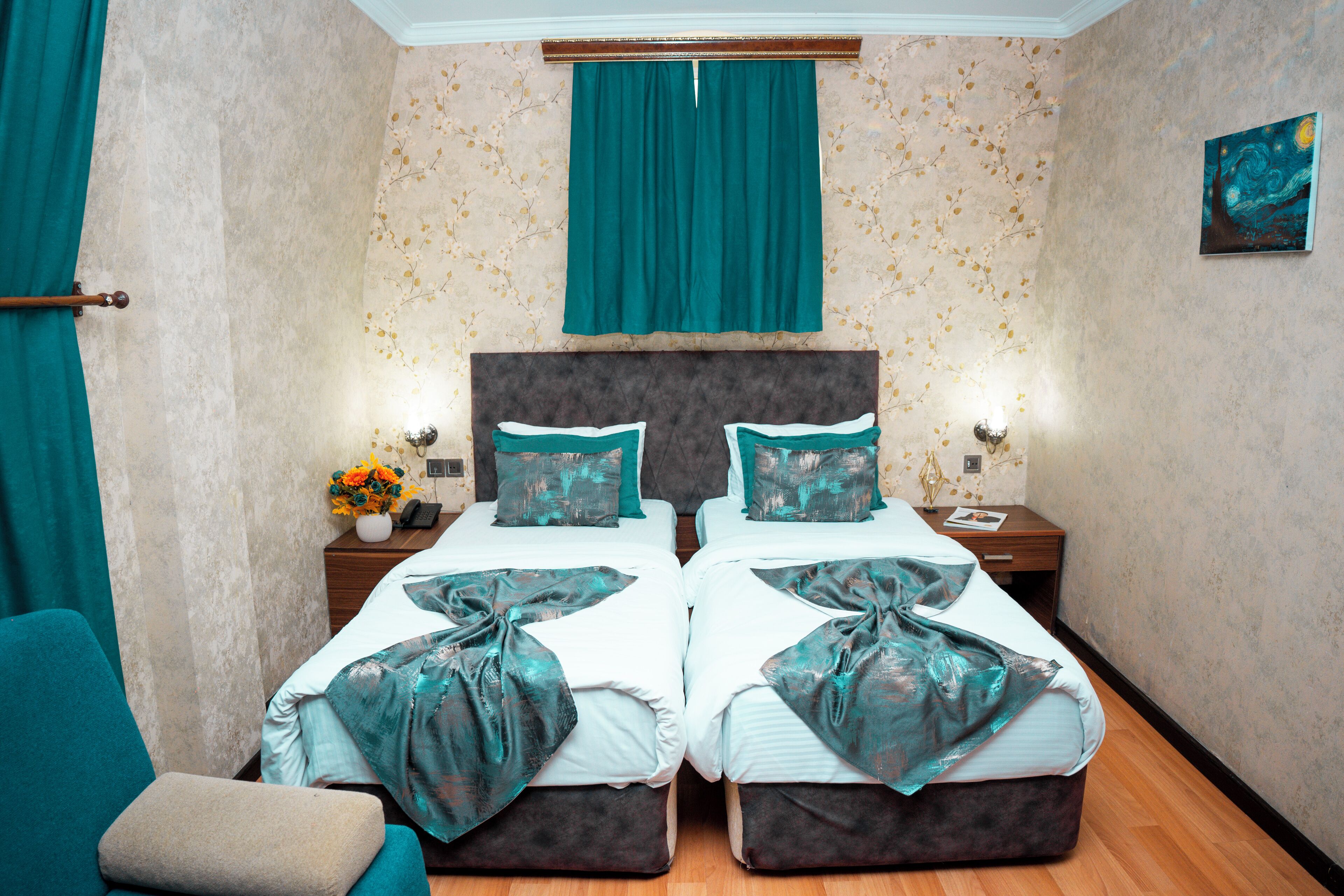 A double room with two beds, a nightstand with a lamp, a small table with a vase of flowers, and a painting on the wall.