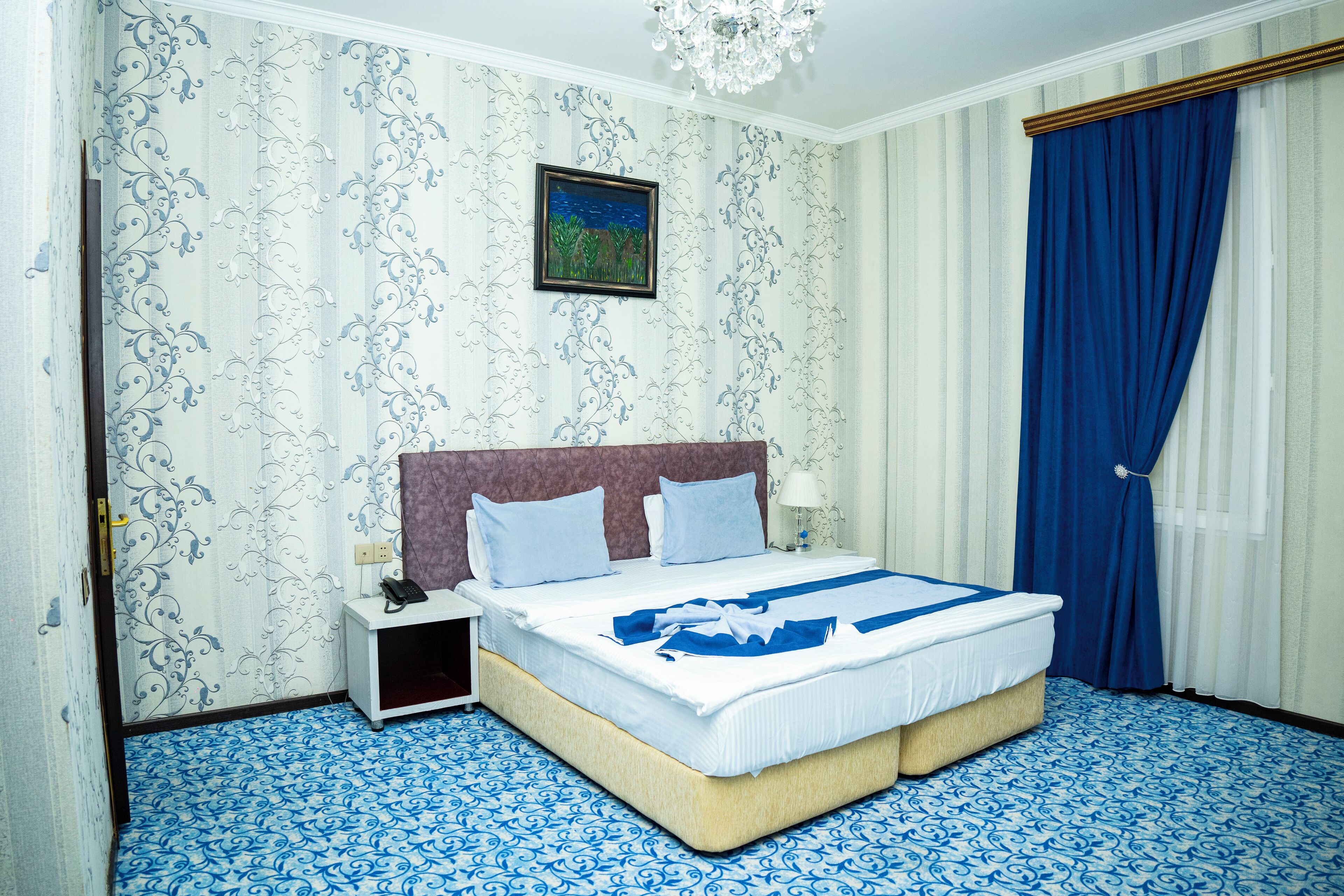 A hotel room with a large bed, a nightstand, a painting on the wall, and a blue curtain.
