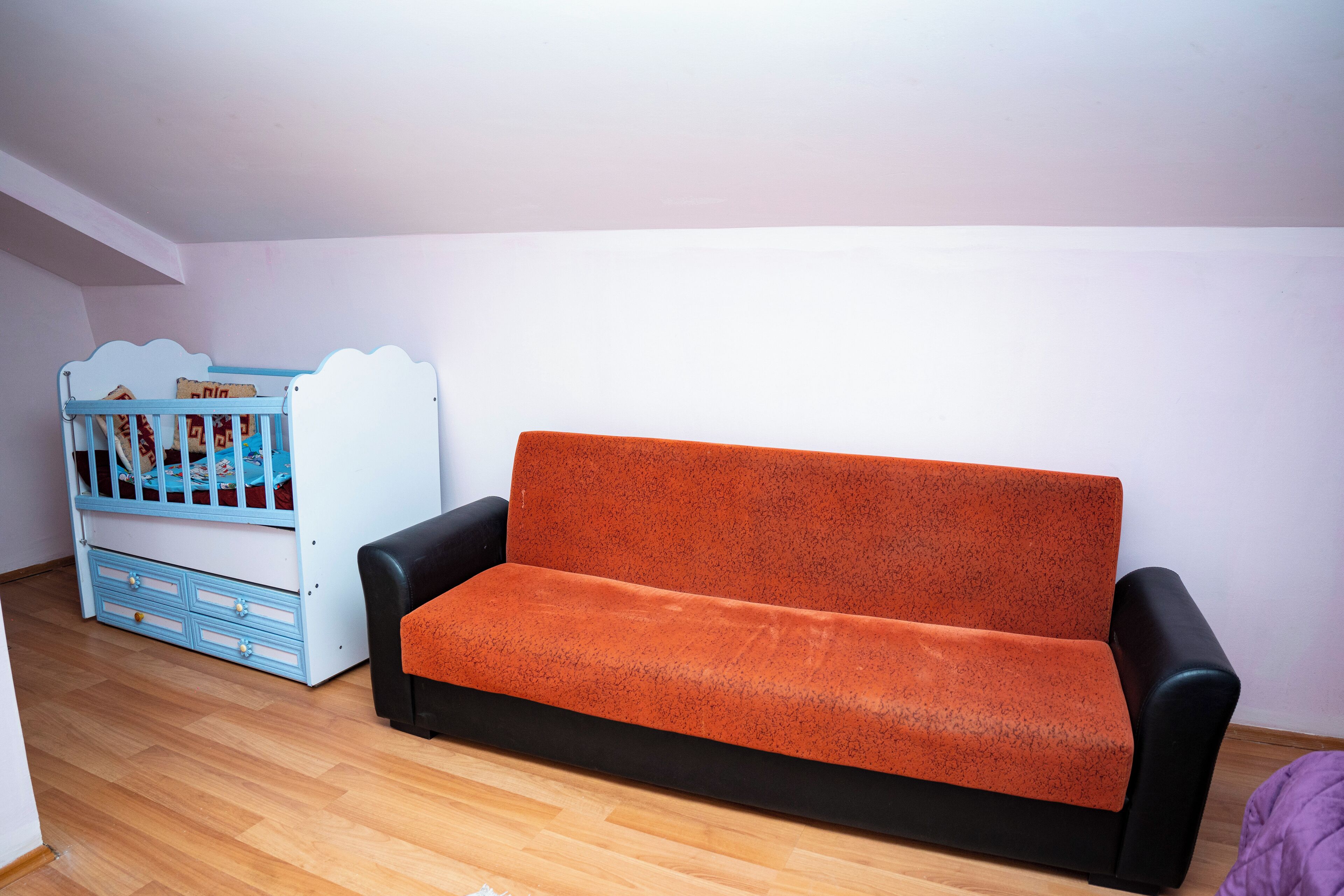 A room with a black and orange sofa, a blue crib, and wooden flooring.