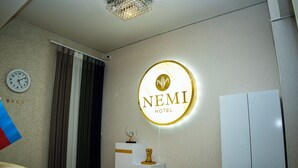 Una camera d'hotel con una sveglia, il cartello NEMI Hotel, un trofeo su un piedistallo e un armadio.