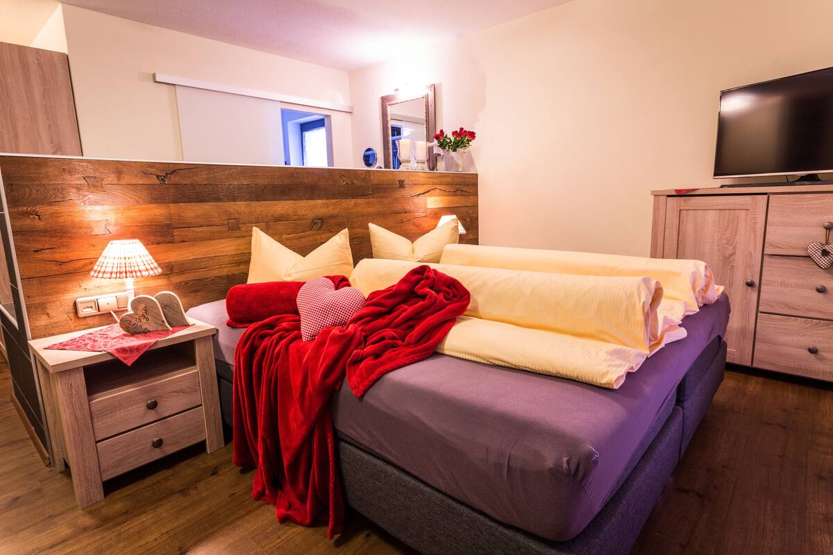 Quarto com cabeceira de madeira, cama com manta vermelha, mesa de cabeceira com abajur e televisão em um armário de madeira.