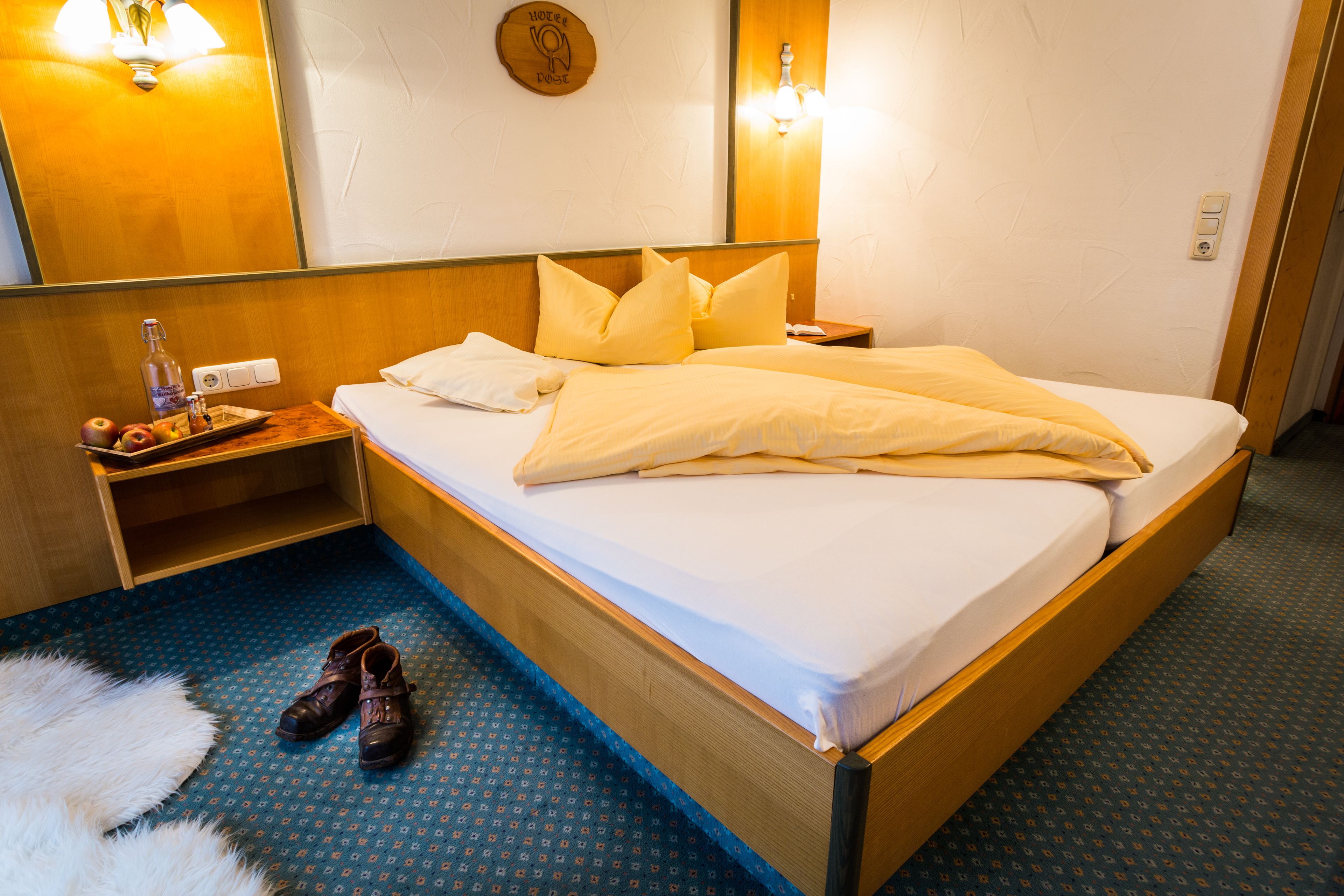 A neatly made bed with pillows, a bedside table with a bottle and glasses, and a pair of shoes on the floor.