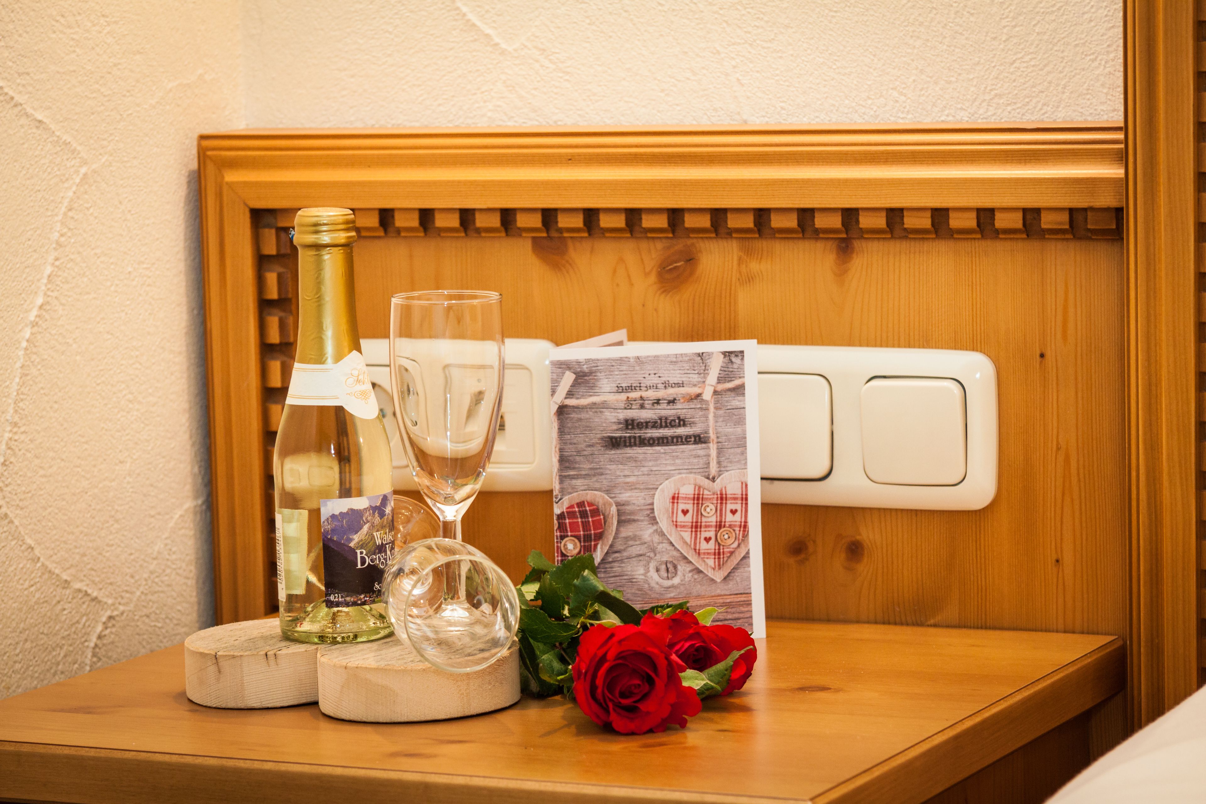 Une table de chevet en bois sur laquelle se trouvent une bouteille de vin, un verre, un bol décoratif, une carte et une rose rouge.