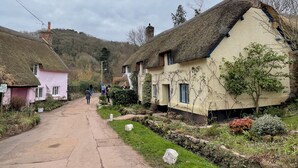 A narrow village street lined with traditional thatched-roof cottages, a clear sky, and a sign for a local cafe.