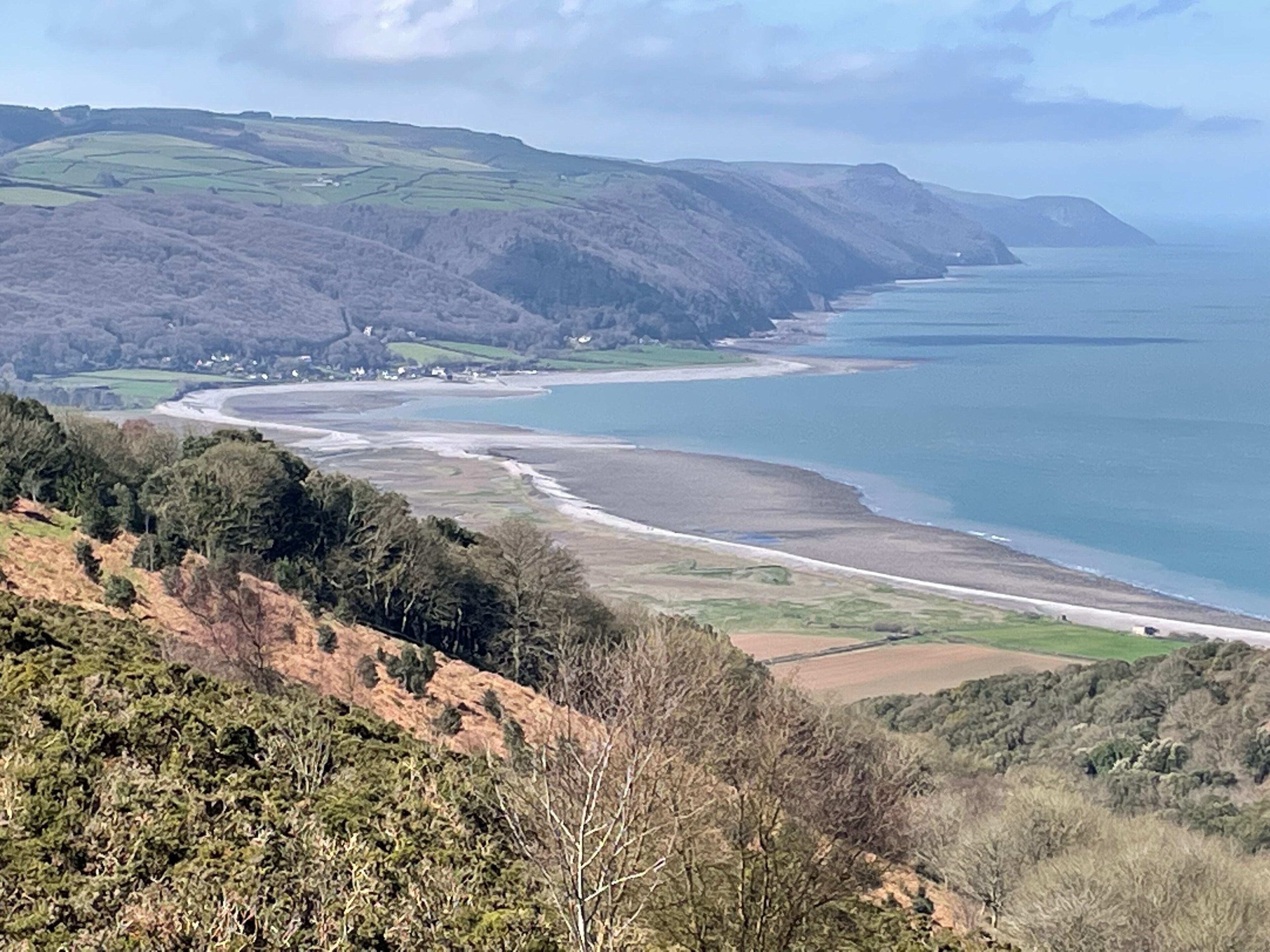 A coastal landscape with a beach, rolling hills, and a distant view of the sea.