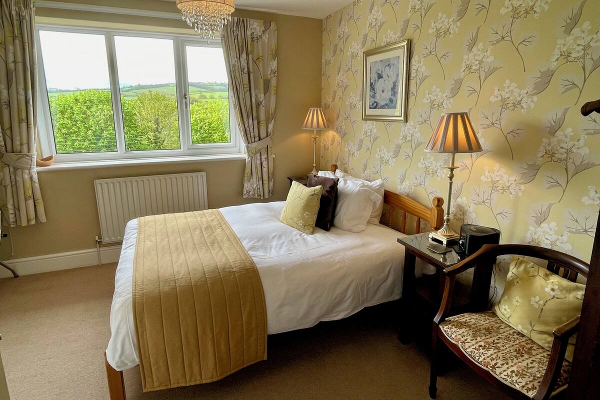 A bedroom with a bed, a chair, a radiator, a window with curtains, and a chandelier.