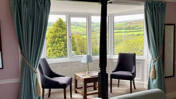 A bedroom with a four-poster bed, two chairs, a small table with a lamp, and a view of green fields through large windows.
