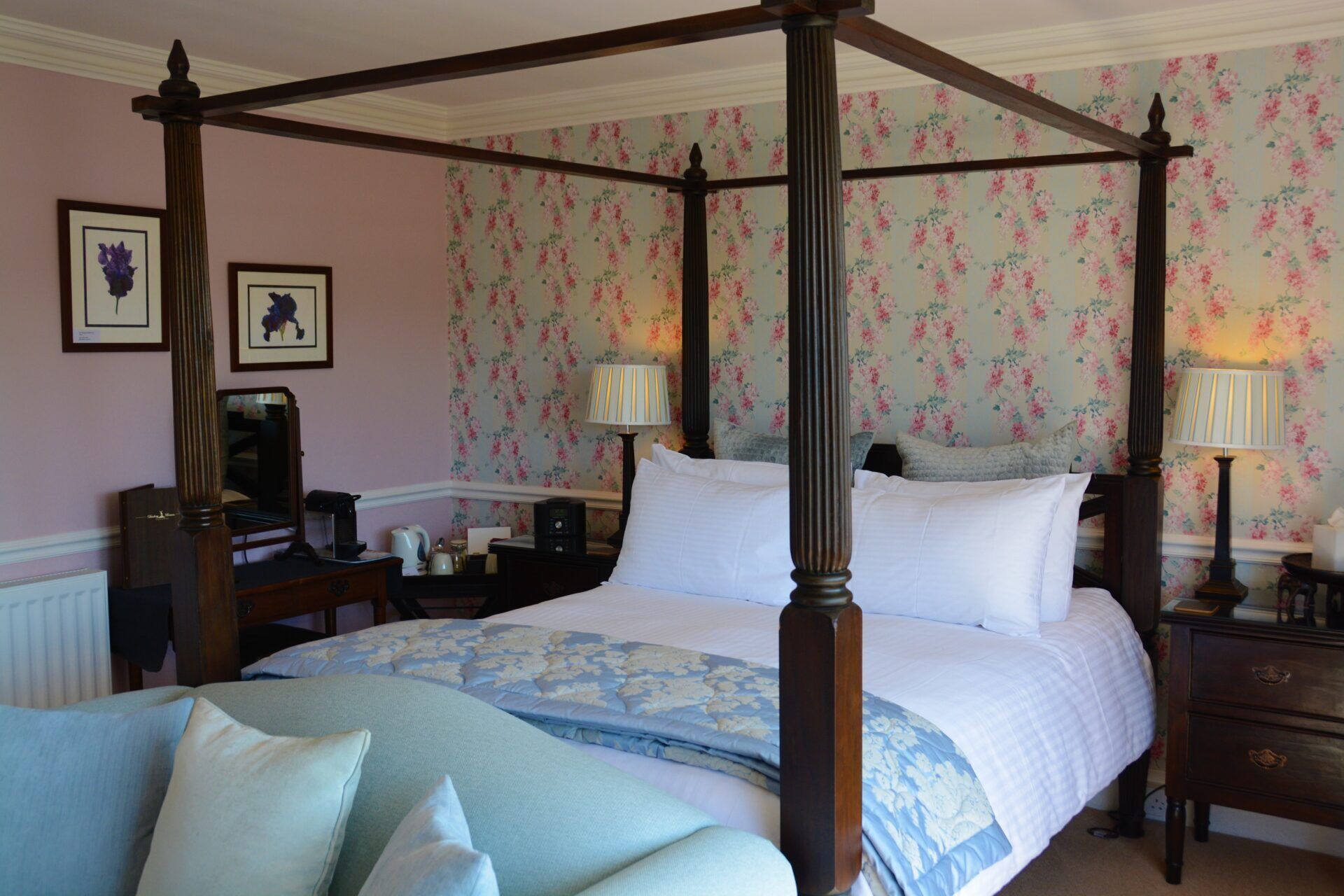 A four-poster bed with a floral wallpaper background, a bedside table with a lamp, and a nightstand with a lamp.