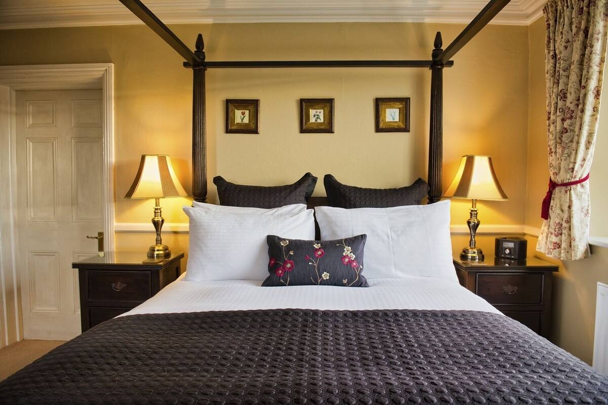A four-poster bed with a dark textured bedspread, two matching pillows, and a single decorative pillow with floral embroidery.
