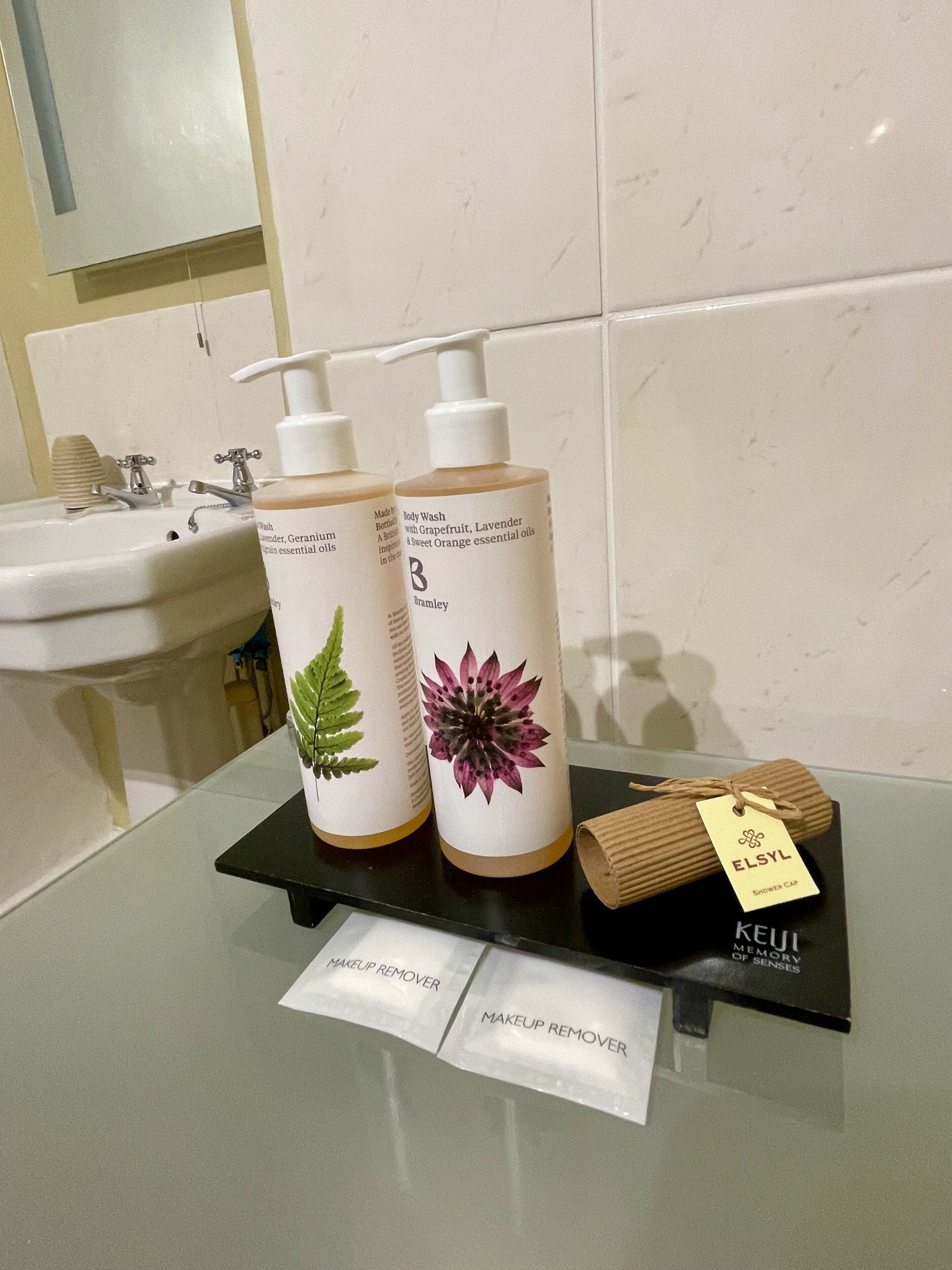 Casa de banho com uma pia, duas embalagens de gel de banho, um tapete de banho de madeira e um tabuleiro preto com uma toalha e uma etiqueta.