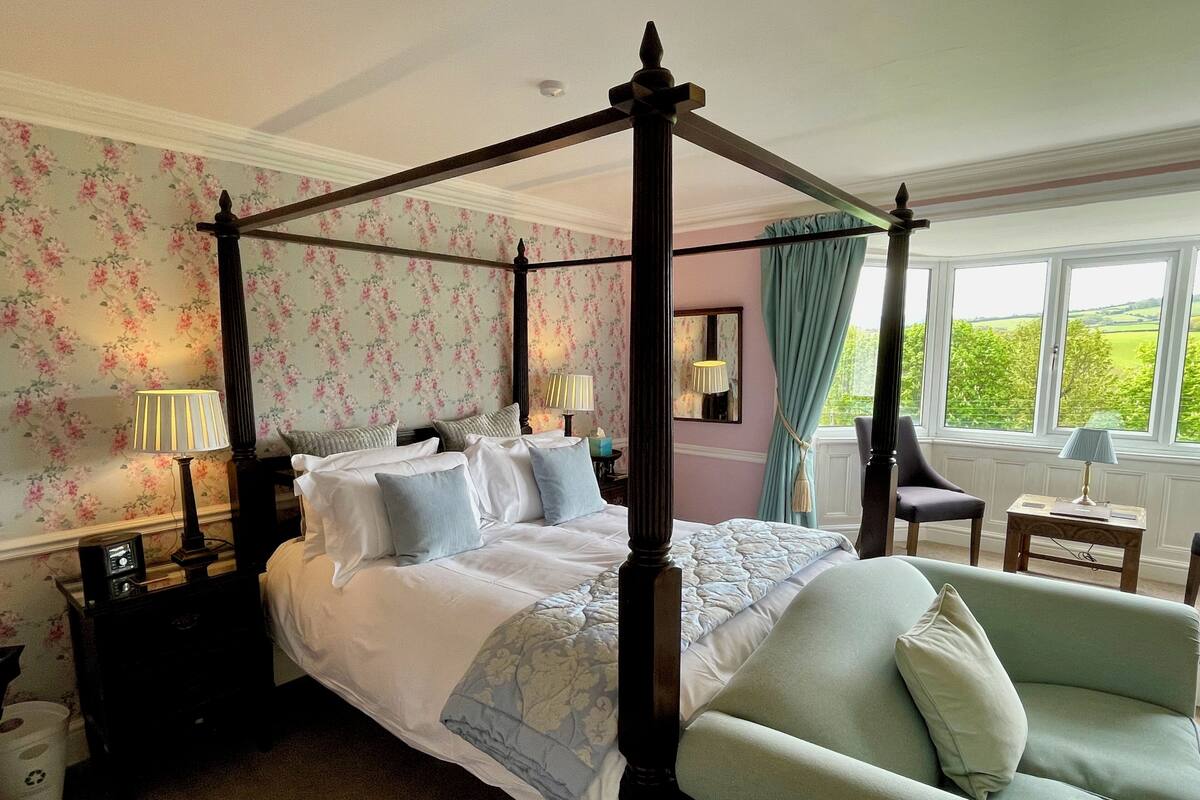 A four-poster bed with a canopy, a sitting chair, a bedside table with a lamp, and a window with a view of greenery.