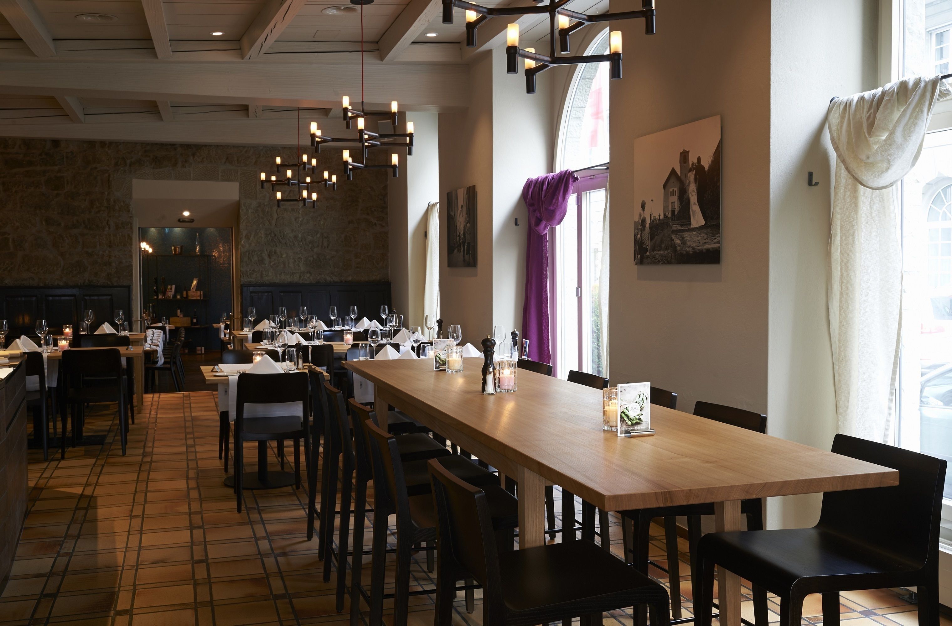 Restaurante com uma longa mesa de madeira, várias cadeiras e luminárias pendentes.