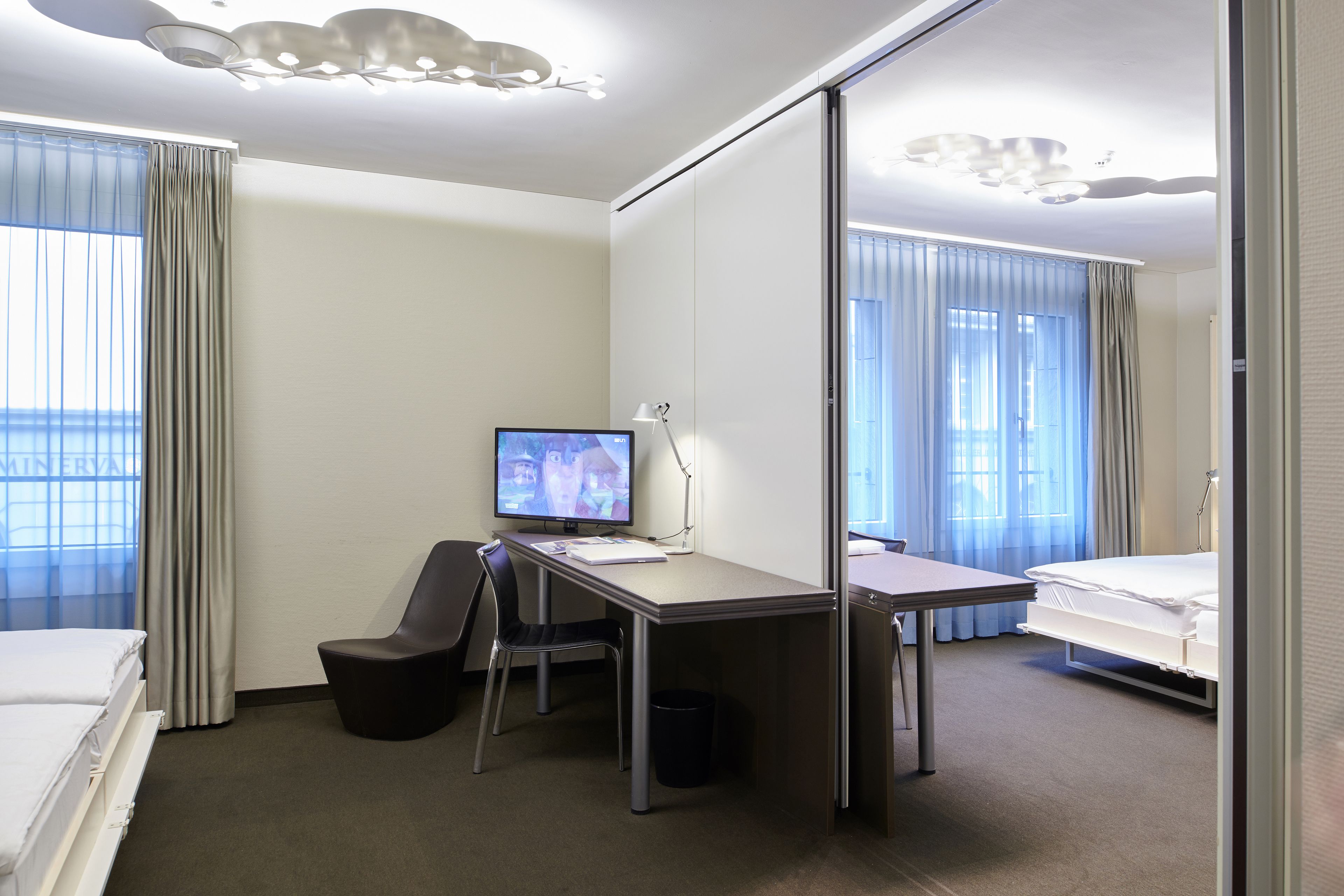 A hotel room with a bed, desk, chair, TV, and large windows with curtains.
