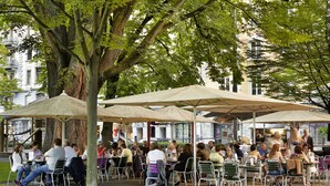 An outdoor café with tables and chairs under large umbrellas, surrounded by trees and buildings. - Hotel Continental Park (Lucerne)