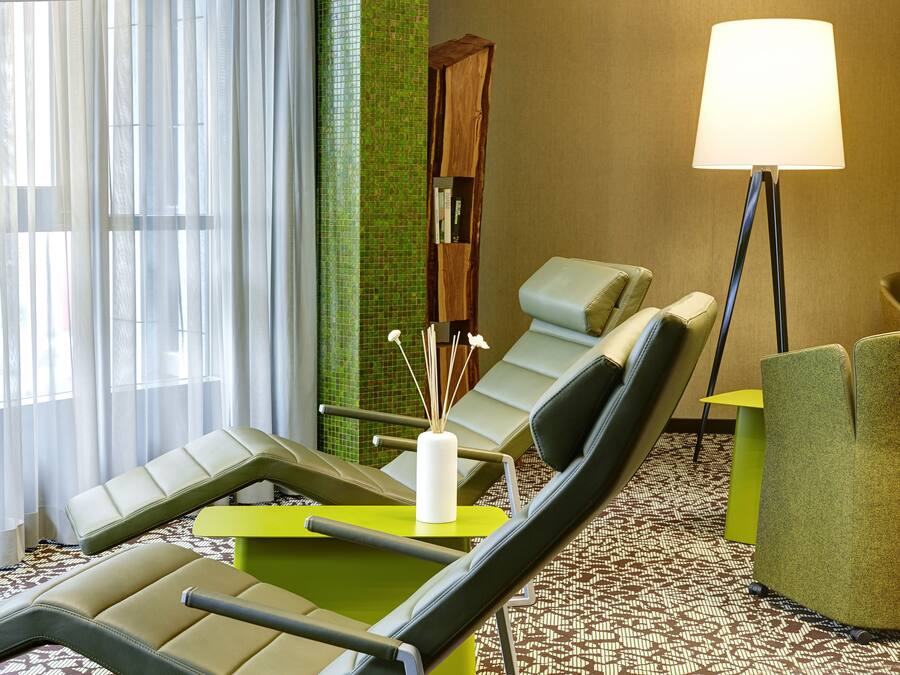 Habitación de hotel con zona de estar que incluye dos sillones, una mesita con un jarrón, una estantería y una lámpara de pie.