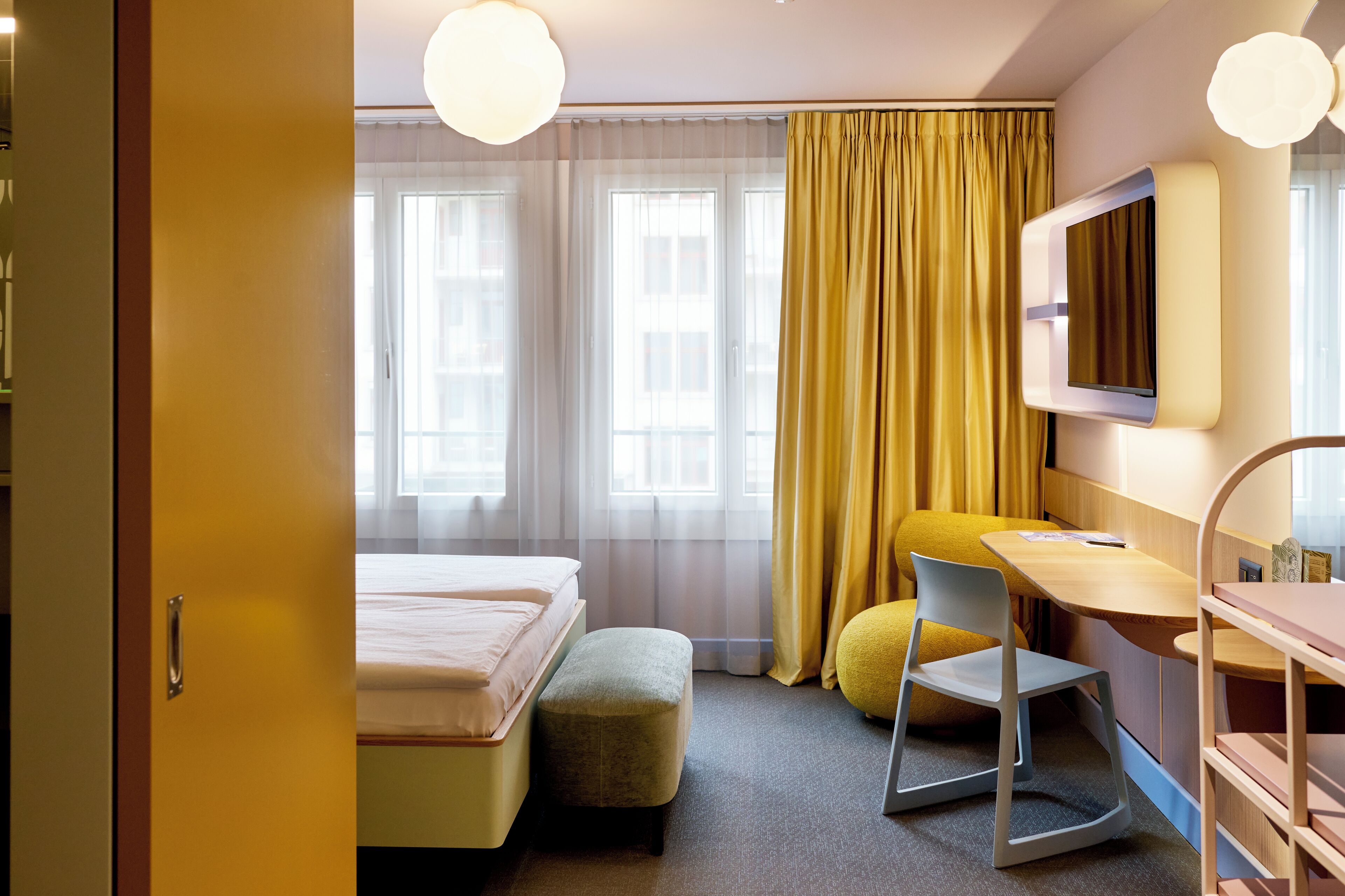 Quarto de hotel com cama, escrivaninha, cadeira, televisão e janela com cortinas.