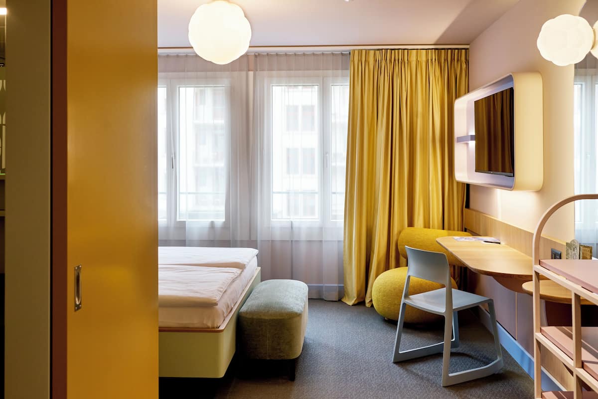 A hotel room with a bed, a desk, a chair, a TV, and a window with curtains.