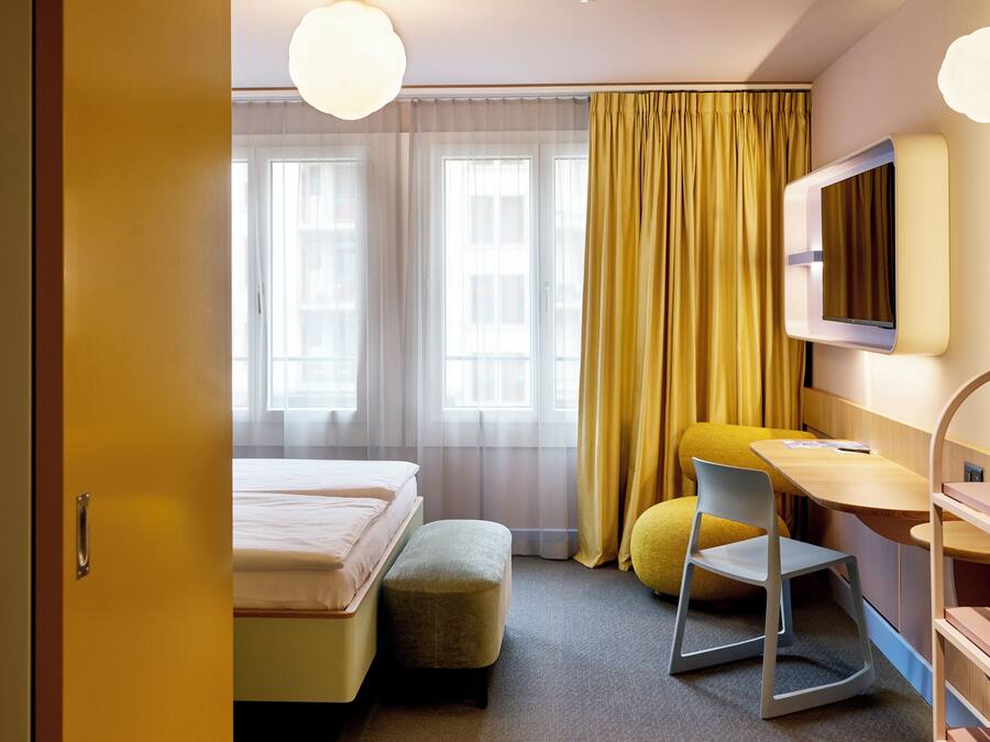 A hotel room with a bed, a desk, a chair, a TV, and a window with curtains.