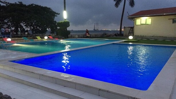 A swimming pool with two sections, surrounded by lounge chairs and palm trees, with a view of a distant building. - Fini Hotel Bobende (Limbe)