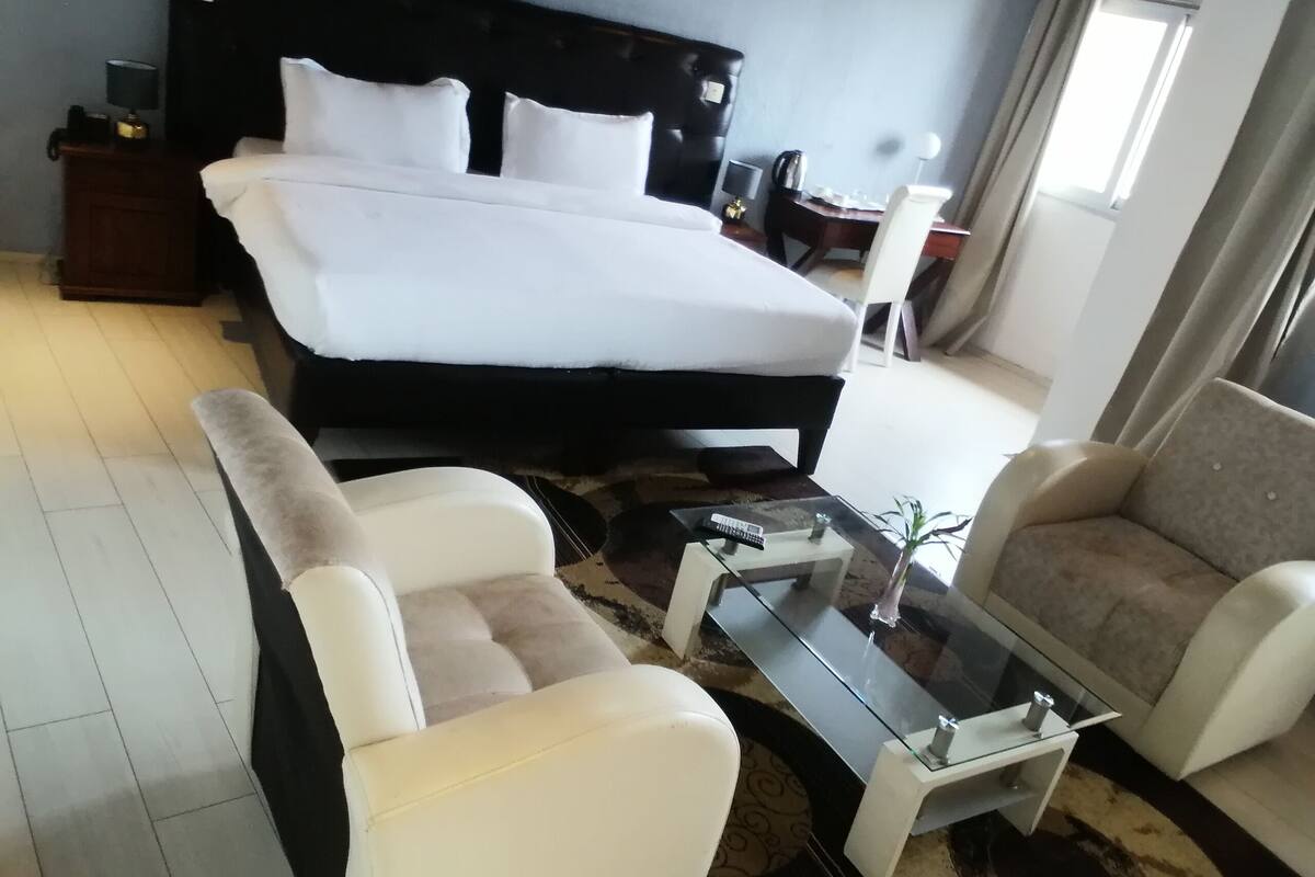 Una habitación de hotel con una cama grande, dos sillones, una mesa de centro de cristal, una mesita de noche y una lámpara.