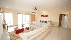 A spacious living room with a large beige sectional sofa, a red coffee table, and a ceiling fan. - Fini Hotel Bobende (Limbe)