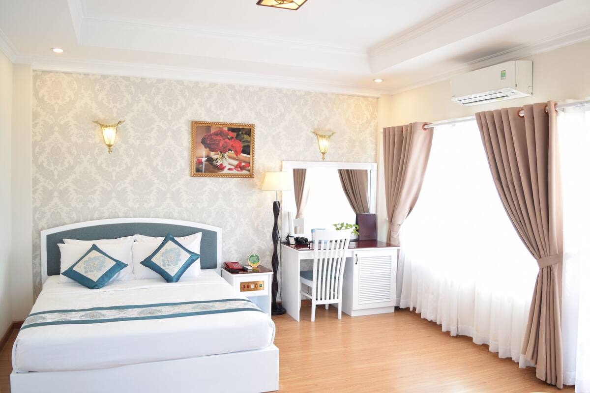 Quarto de hotel com cama, escrivaninha, cadeira, luminária e cortinas.