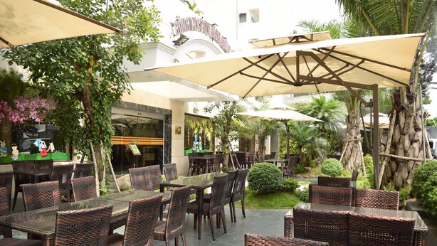 An outdoor dining area with wicker furniture and large umbrellas, surrounded by greenery and a building facade. - Rang Dong Hotel (Ho Chi Minh City)