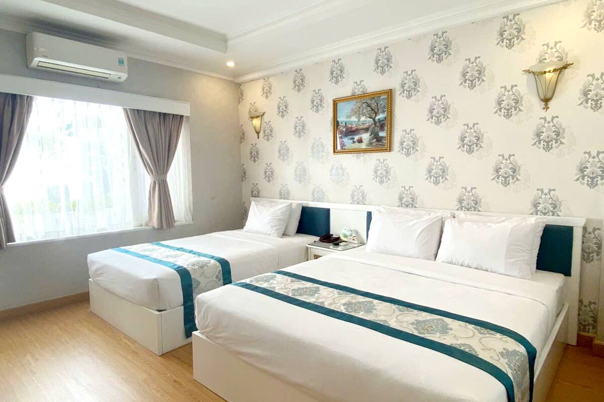 Quarto de hotel com duas camas, janela com cortinas, aparelho de ar condicionado, mesa de cabeceira com abajur e papel de parede estampado.