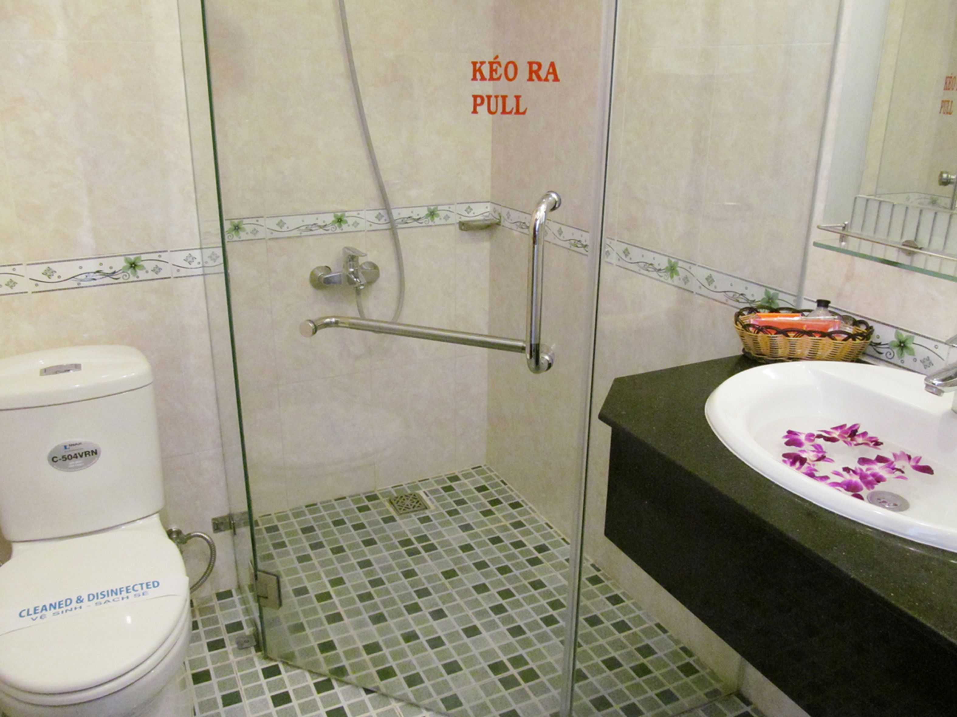 A bathroom with a toilet, a shower with a pull handle, a sink with a decorative flower, and a basket on the counter.