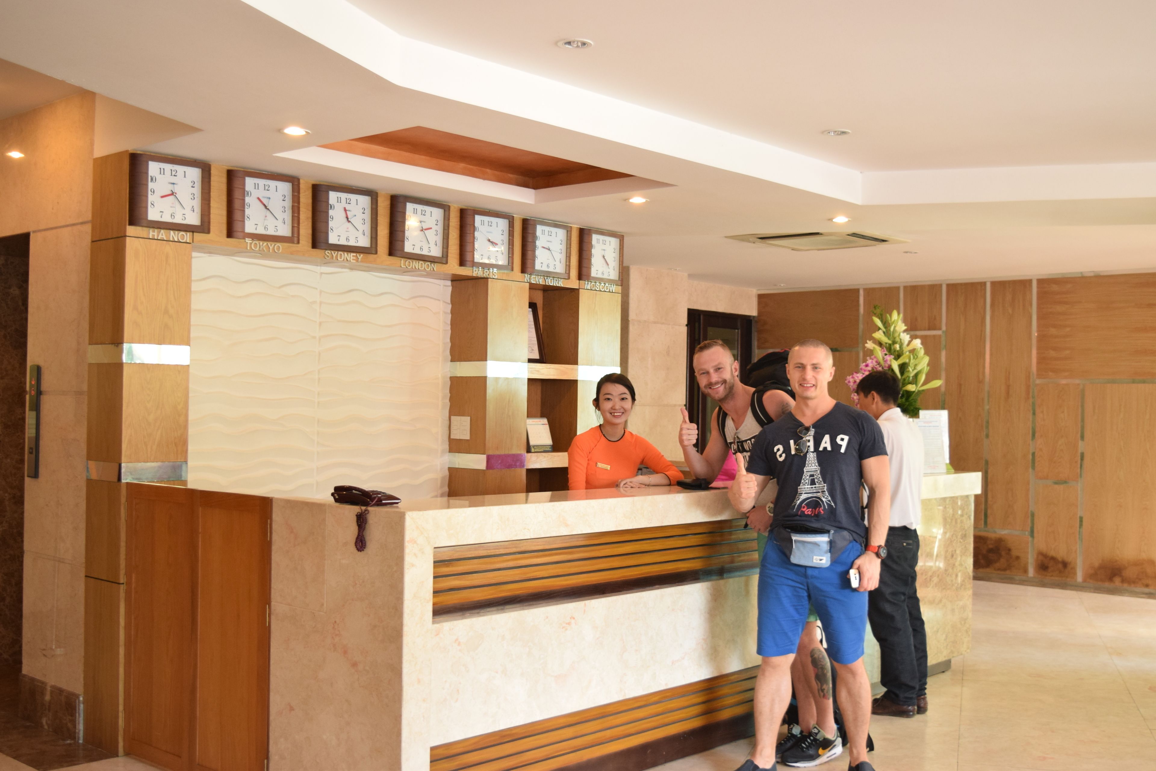 Una recepción con relojes mostrando horarios de diferentes ciudades, una recepcionista y dos huéspedes.