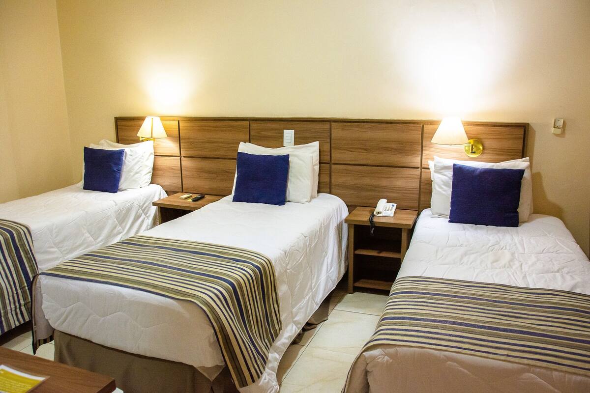 Una habitación de hotel con tres camas individuales, cada una con cabecera y mesitas de noche de madera.