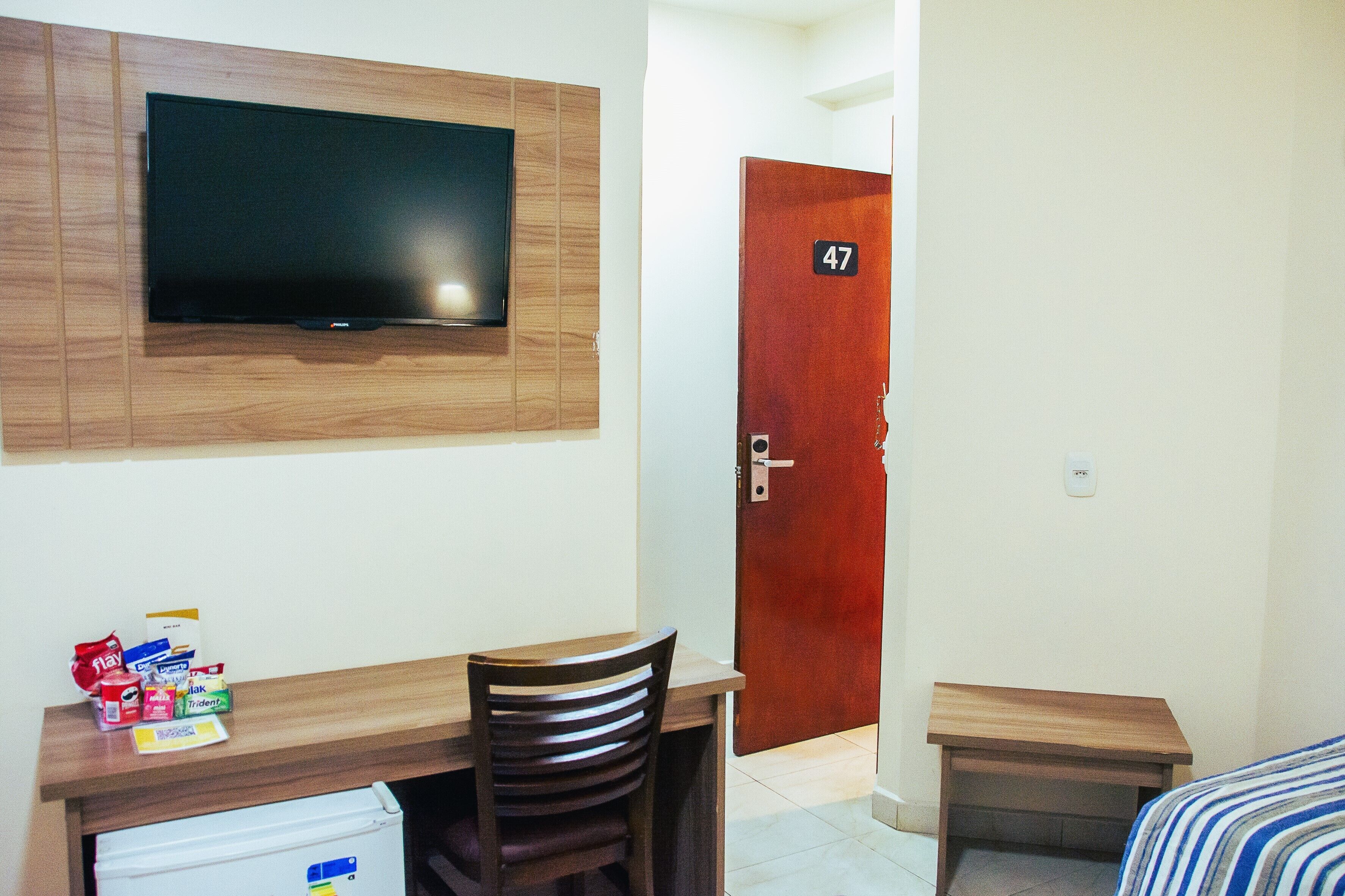 A hotel room with a TV mounted on the wall, a wooden desk with a chair, a small bedside table, and a door with the number 47.
