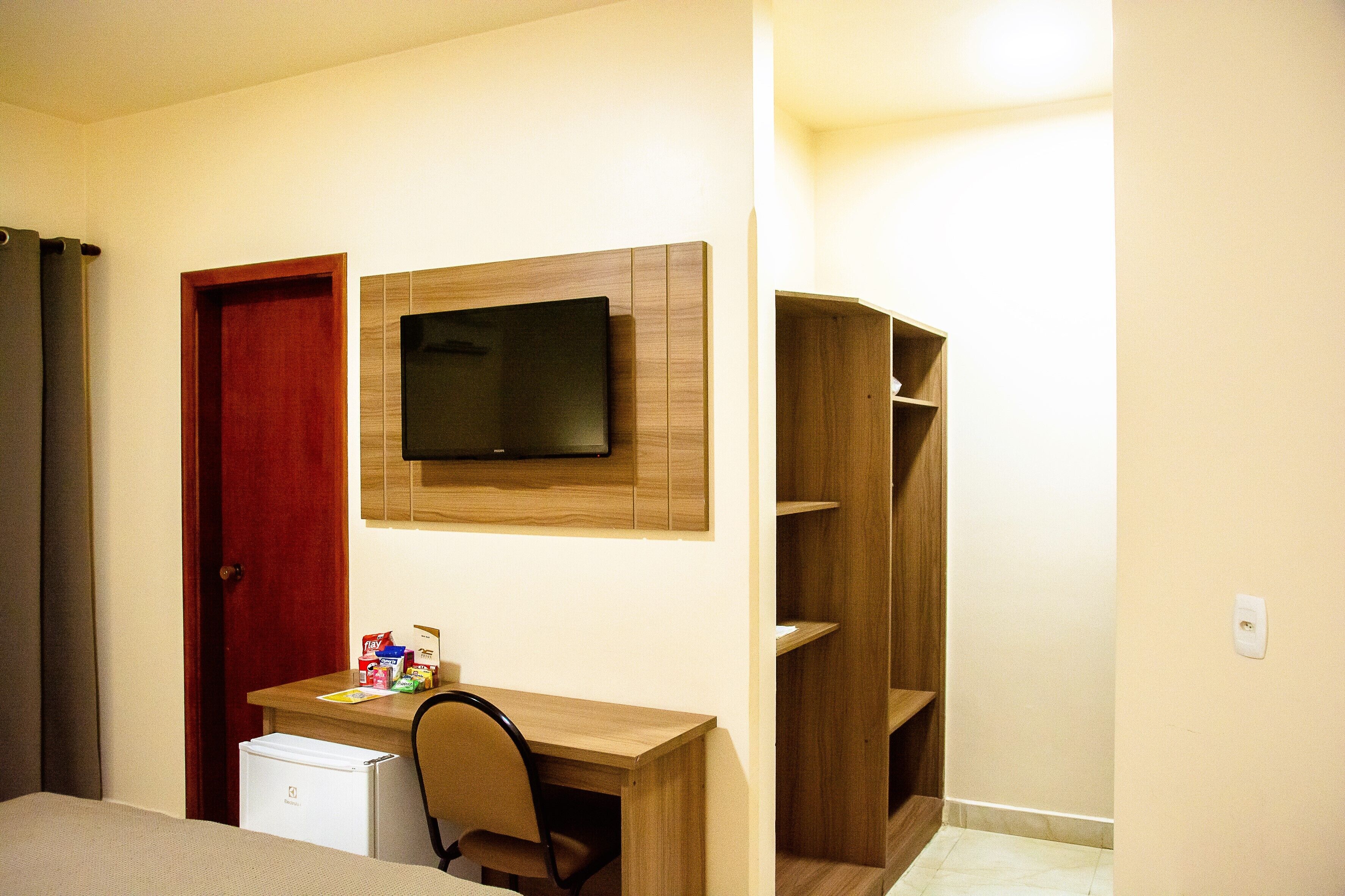 Ein Zimmer mit einem Holztisch, einem an der Wand montierten Fernseher, einer roten Tür und einem kleinen weißen Kühlschrank.