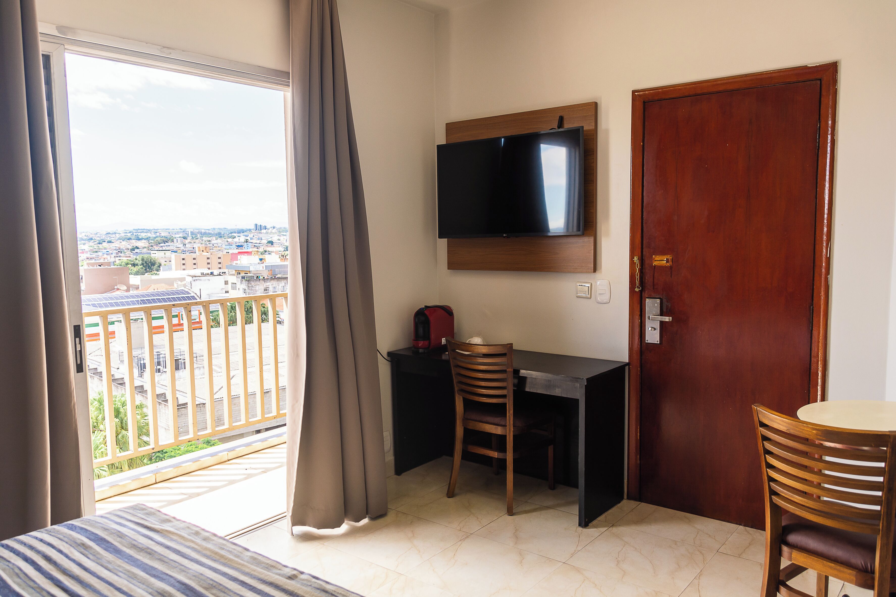 A room with a TV mounted on the wall, a wooden door, a desk with a chair, and a view of the city through a balcony.