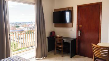 A room with a TV mounted on the wall, a wooden door, a desk with a chair, and a view of the city through a balcony.