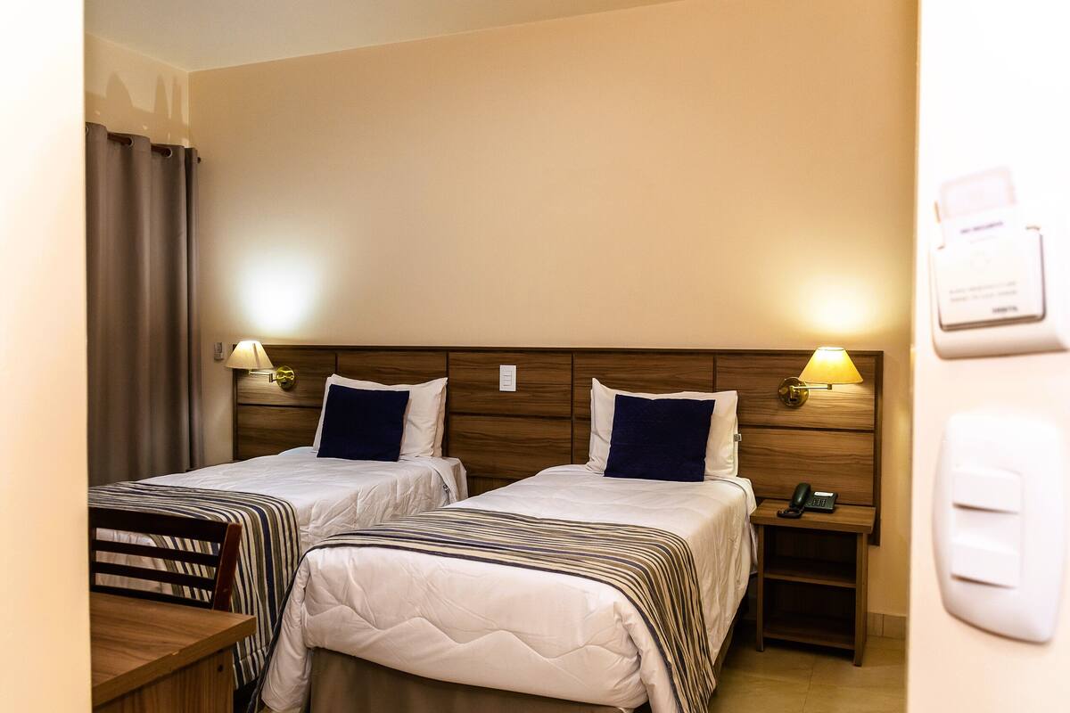 Habitación de hotel con dos camas, lámparas de noche, una mesita de madera, un teléfono y un interruptor de luz en la pared.
