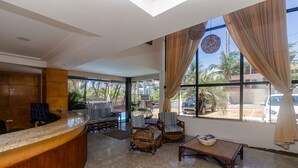 A reception area with a curved wooden counter, wicker chairs, a small table with a bowl, and large windows with curtains. - Apart Hotel Vespucci Residence (Natal)