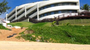 A modern multi-story building with a curved facade, a flat roof, and a landscaped area with grass and plants in front. - Apart Hotel Vespucci Residence (Natal)